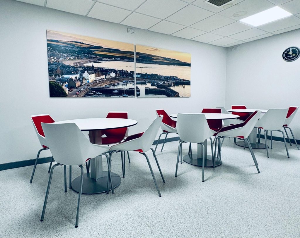 NOV catering area with white tables, red chairs and print of Stonehaven harbour.